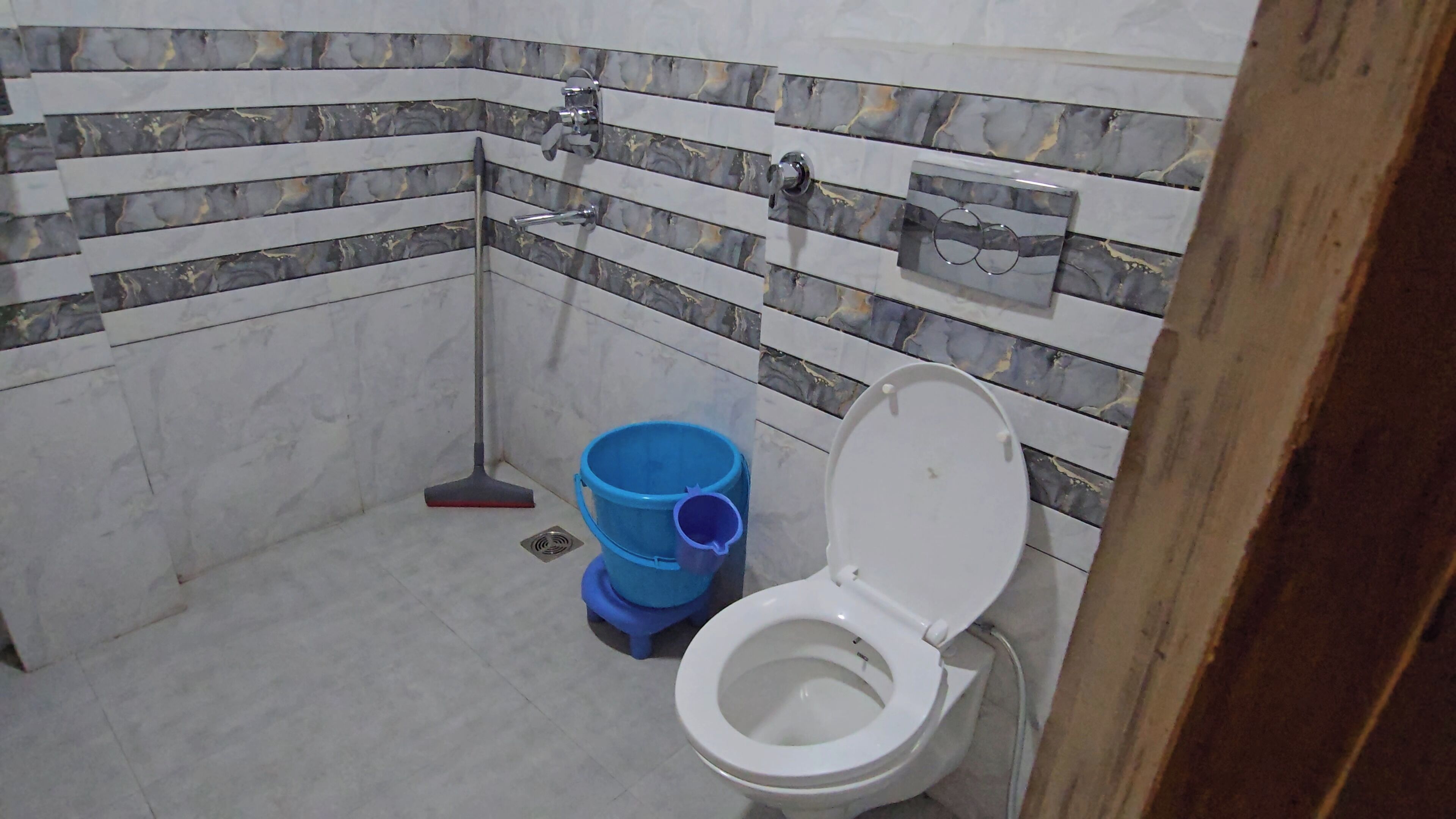 A bathroom with a toilet, a blue bucket, and a broom.