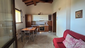 A compact kitchen with wooden cabinets, a dining table with chairs, and a red sofa with floral cushions. - Val D'Orcia Window. Panoramic balcony. Pool. (Contignano)