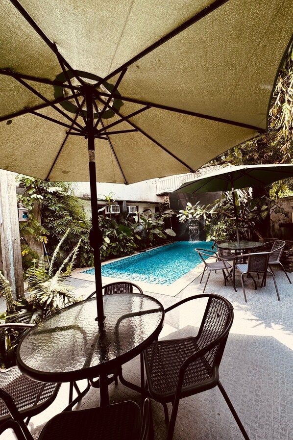 Un espace piscine extérieur doté d’une table et de chaises en verre, installé sous une pergola, et entouré d’une végétation luxuriante ainsi que d’une cascade.
