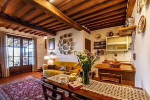Un salon chaleureux avec un plafond en bois, un canapé jaune, une table à manger recouverte d’une nappe en dentelle et un coin cuisine doté d’un plan de travail en bois.