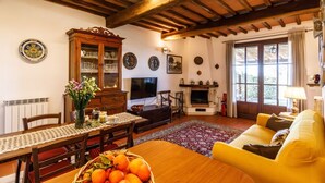 A cozy living room with a wooden ceiling, a dining table with chairs, a sofa, a fireplace, and a television. - GuestHost - Nestled between the Crete Senesi and Chianti, this home offers a breathtaking view of the countryside and Siena, just a 10-minute drive away. Part of a renovated historic farmhouse, it is a well-furnished townhouse with a private garden and v (Castelnuovo Bardenga)