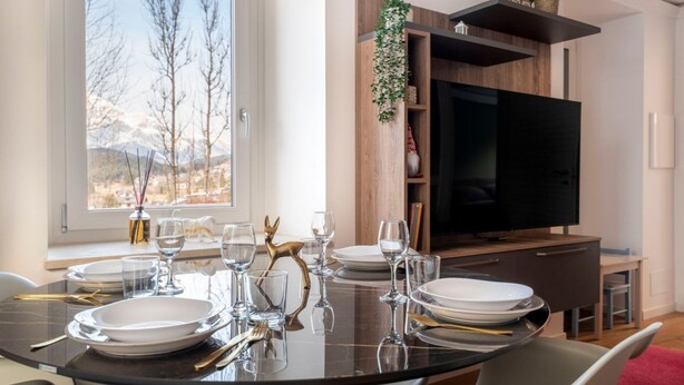 Comedor con una mesa redonda dispuesta para cuatro personas, un televisor de pantalla plana y una estantería con una planta.