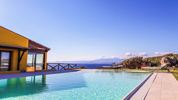 A pool area with clear blue water, a yellow building, and a view of the sea and mountains.