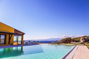 Une piscine à l’eau d’un bleu cristallin, un bâtiment jaune et une vue sur la mer et les montagnes.