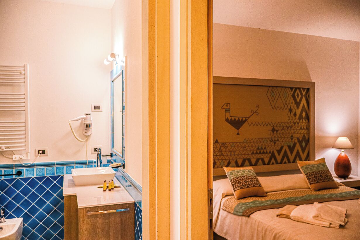 A hotel room with a bathroom featuring a sink, mirror, and towel rack. The room has a bed with pillows and a bedside table with a lamp.