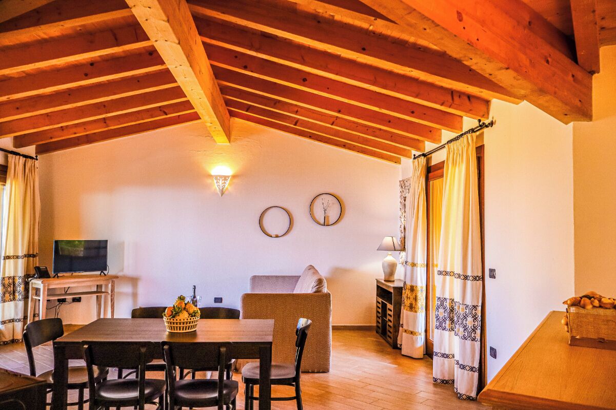 A cozy room with a wooden ceiling, a dining table with chairs, a sofa, a TV, and a wall clock.