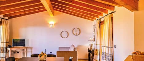 A cozy room with a wooden ceiling, a dining table with chairs, a sofa, a TV, and a wall clock.