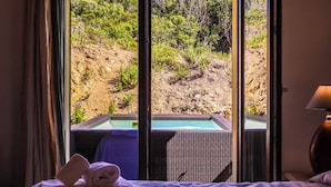 A room with a bed, a window with a view of a pool and natural landscape.
