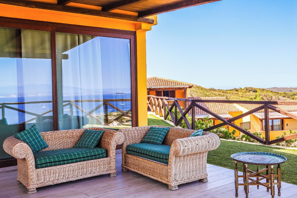 A wicker sofa with green cushions on a wooden deck with a glass door, overlooking a coastal landscape.