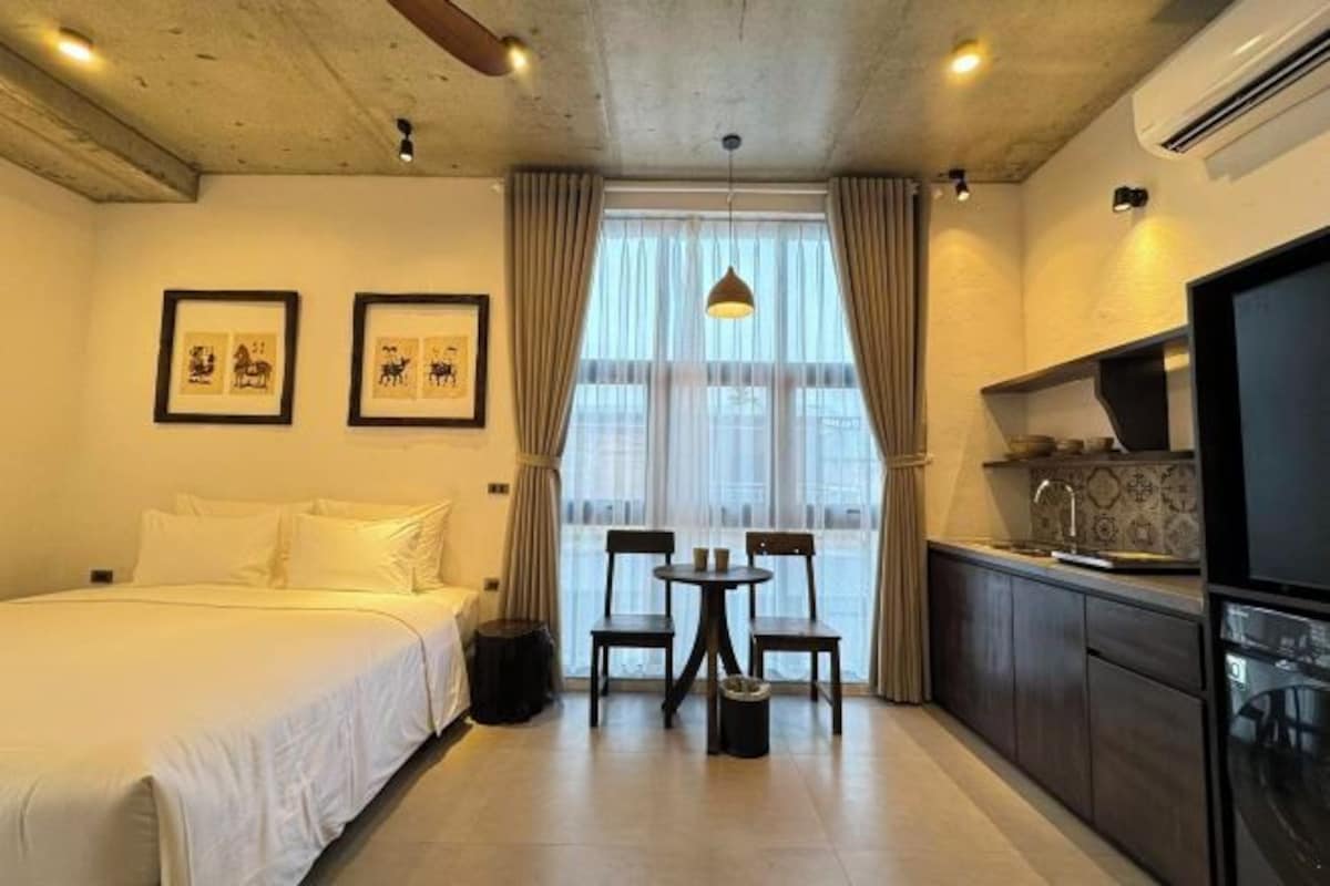 Ett kompakt hotellrum med en säng, ett litet matbord med stolar, ett litet kök och ett stort fönster med gardiner.
