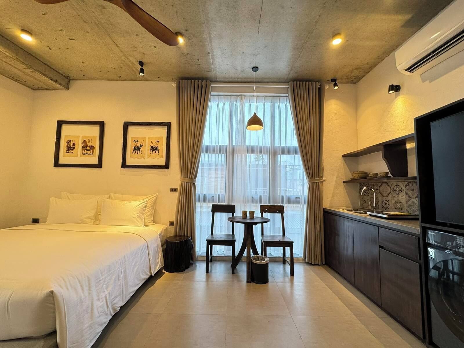 A hotel room with a bed, a dining table with chairs, a kitchen area, and a large window with curtains.