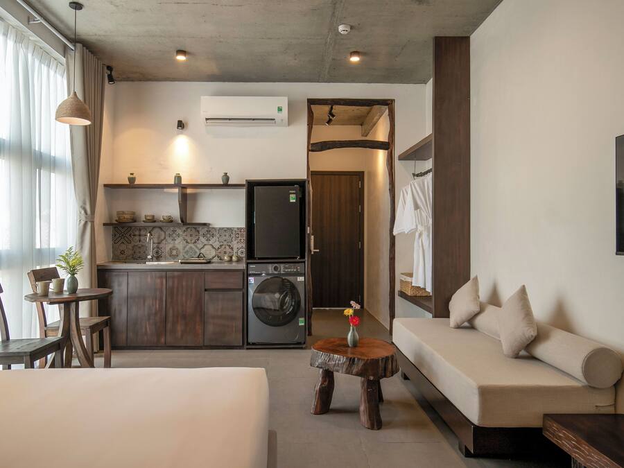 A compact hotel room with a bed, a sofa, a dining table with chairs, a small kitchen area with a washing machine, and an air conditioning unit.