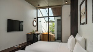 A modern hotel room with a large bed, a flat-screen TV mounted on the wall, a wooden vanity with a sink, and a large window offering a view of the outdoors. - Eco & Rustic Home Hoian (Da Nang)