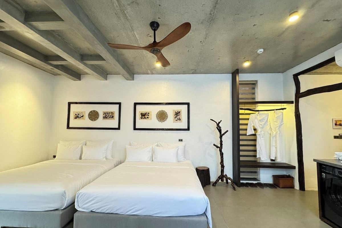 Habitación de hotel con dos camas, ventilador de techo, cuadros en la pared, escalera que lleva a un baño y un mueble negro.