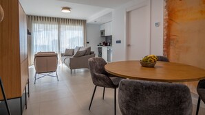 A modern living room with a round dining table, grey upholstered chairs, a sofa, and a kitchen area in the background.