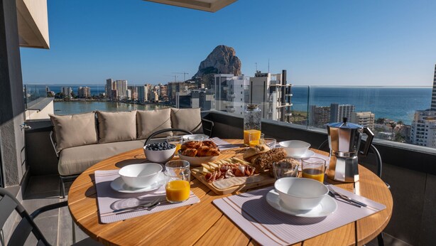 A breakfast table with various food items and drinks, a sofa, and a view of the city and ocean.