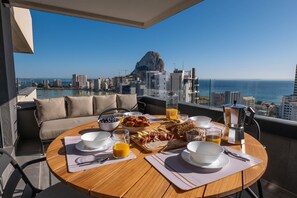 Uma mesa de café da manhã com diversos alimentos e bebidas, um sofá e vista para a cidade e o oceano.