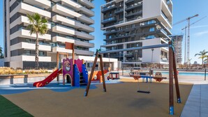 A playground with swings and slides, surrounded by modern apartment buildings and a construction crane in the background.