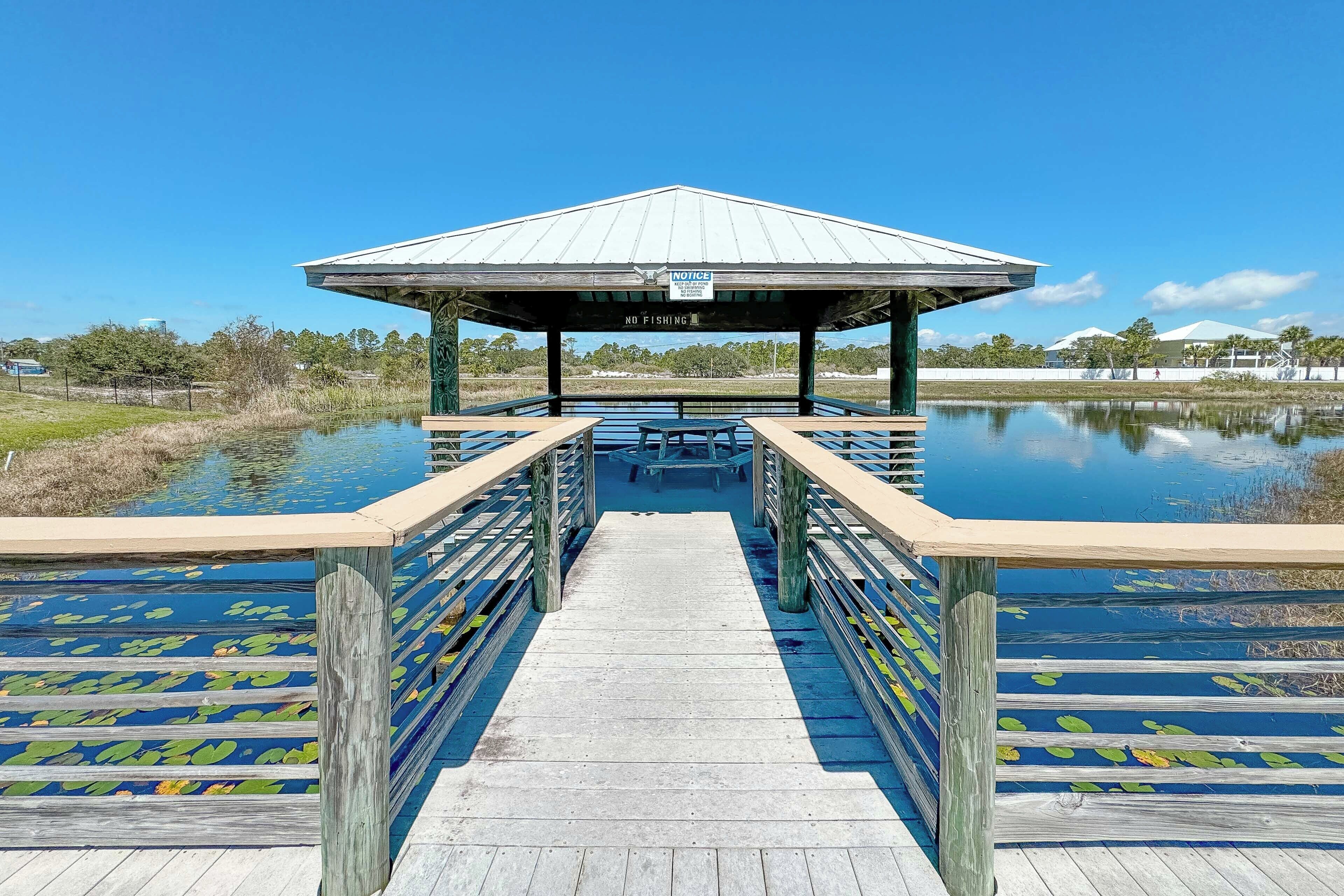 A wooden pier leading to a covered pavilion with a 'NO FISHING' sign, surrounded by water and greenery.