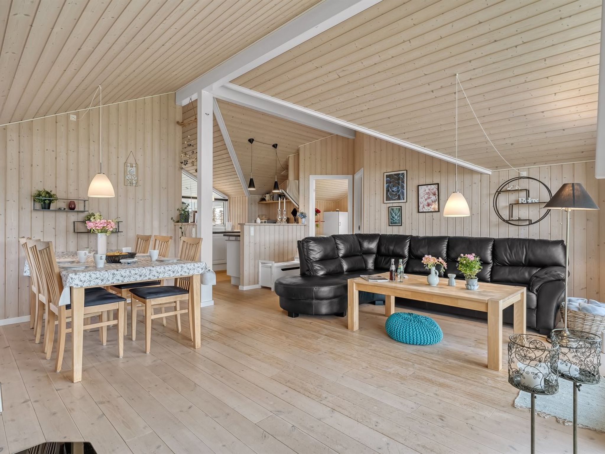 A spacious living area with a wooden ceiling, a black leather sofa, a dining table with chairs, and a kitchen in the background.