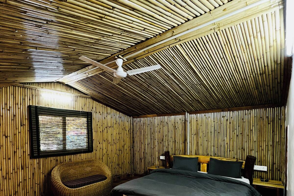 A room with a sloped ceiling, bamboo wall panels, a bed with a dark mattress, a wooden nightstand, a woven chair, and a floral rug.