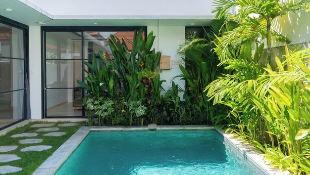 Zona de piscina moderna con una piscina azul cristalina, rodeada de exuberante vegetación tropical y una entrada con puerta de cristal.