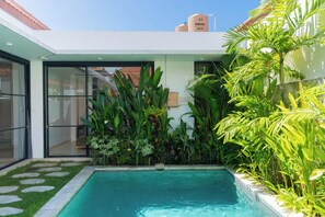 Un espace piscine moderne avec une piscine à l’eau d’un bleu limpide, entourée d’une végétation tropicale luxuriante et doté d’une entrée avec porte en verre.