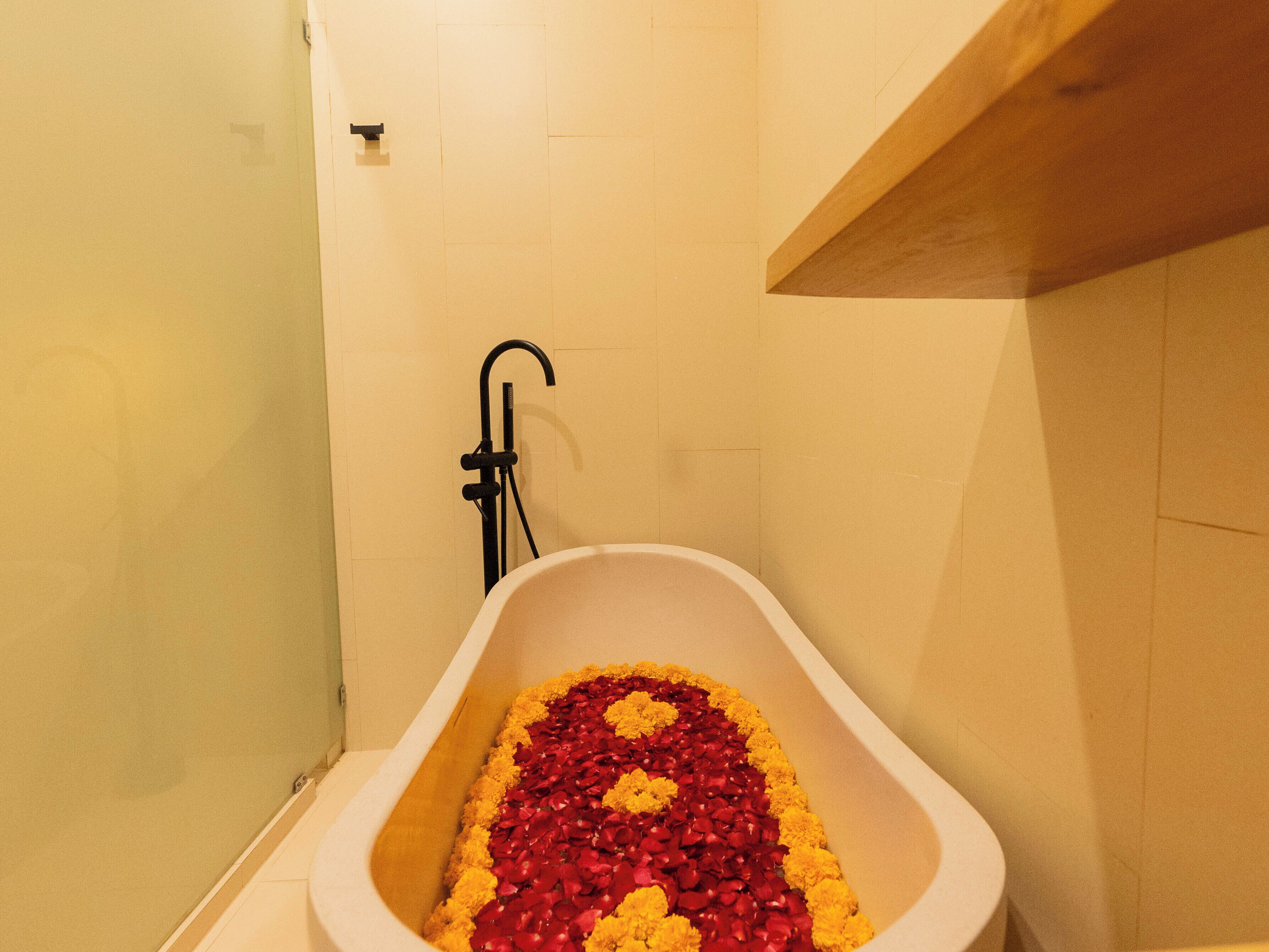 A bathtub filled with rose petals and yellow flowers, with a black faucet and a wooden shelf above.