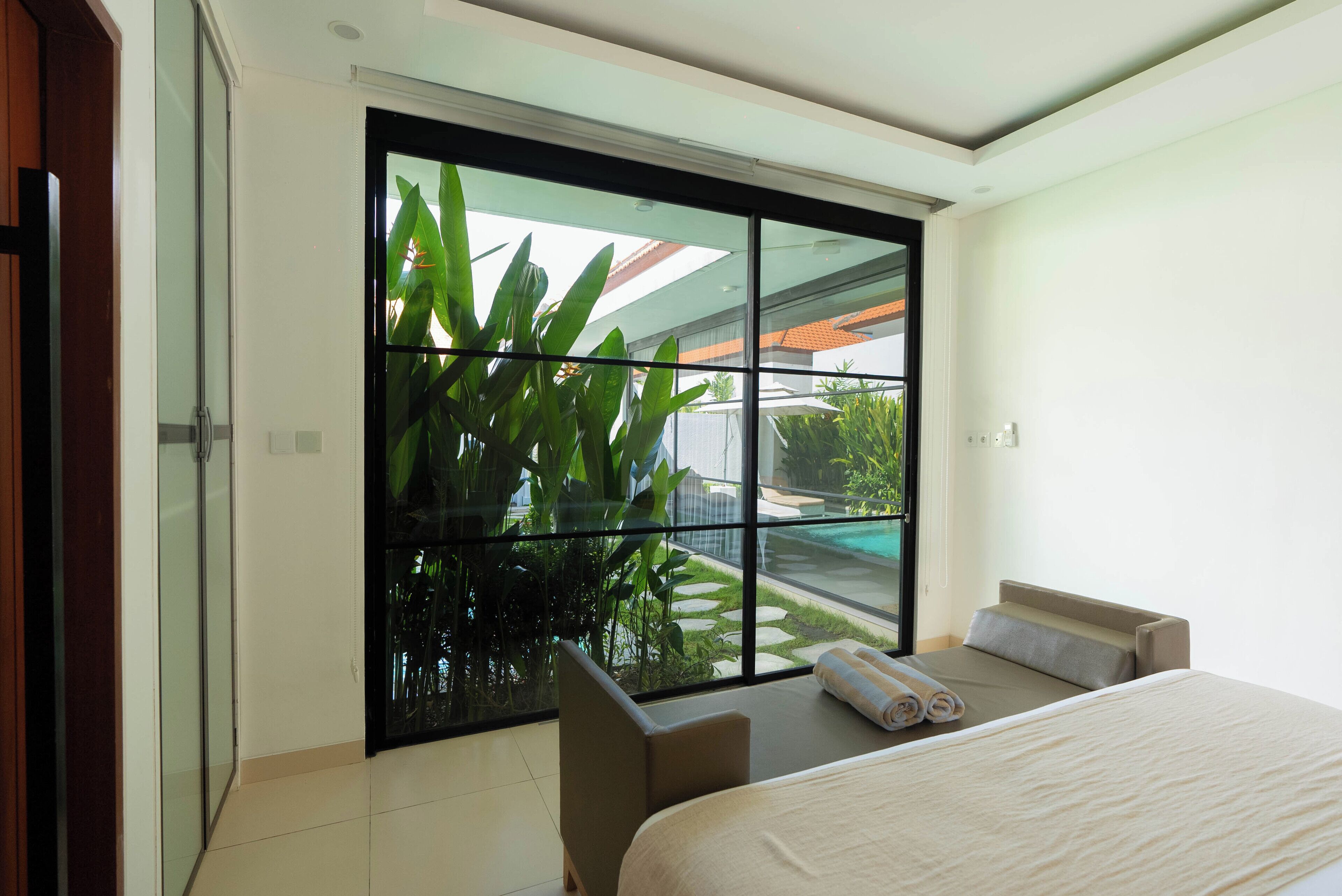 A modern hotel room with a large sliding glass door leading to a pool area, a bed with a neatly made bed, and a small seating area.