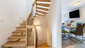 Un interior moderno con una escalera de madera, un comedor con mesa y sillas, y un televisor de pantalla plana montado en la pared.