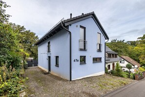 Una casa de dos pisos con fachada azul, un número '47a' visible en la pared, y un pequeño balcón en el piso superior.
