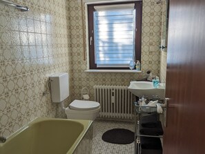 Une salle de bain équipée d’une baignoire, de toilettes, d’un radiateur et d’une fenêtre avec des stores.