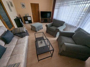 Un salon comprenant un canapé, deux fauteuils, une table basse en verre, un téléviseur et une porte.
