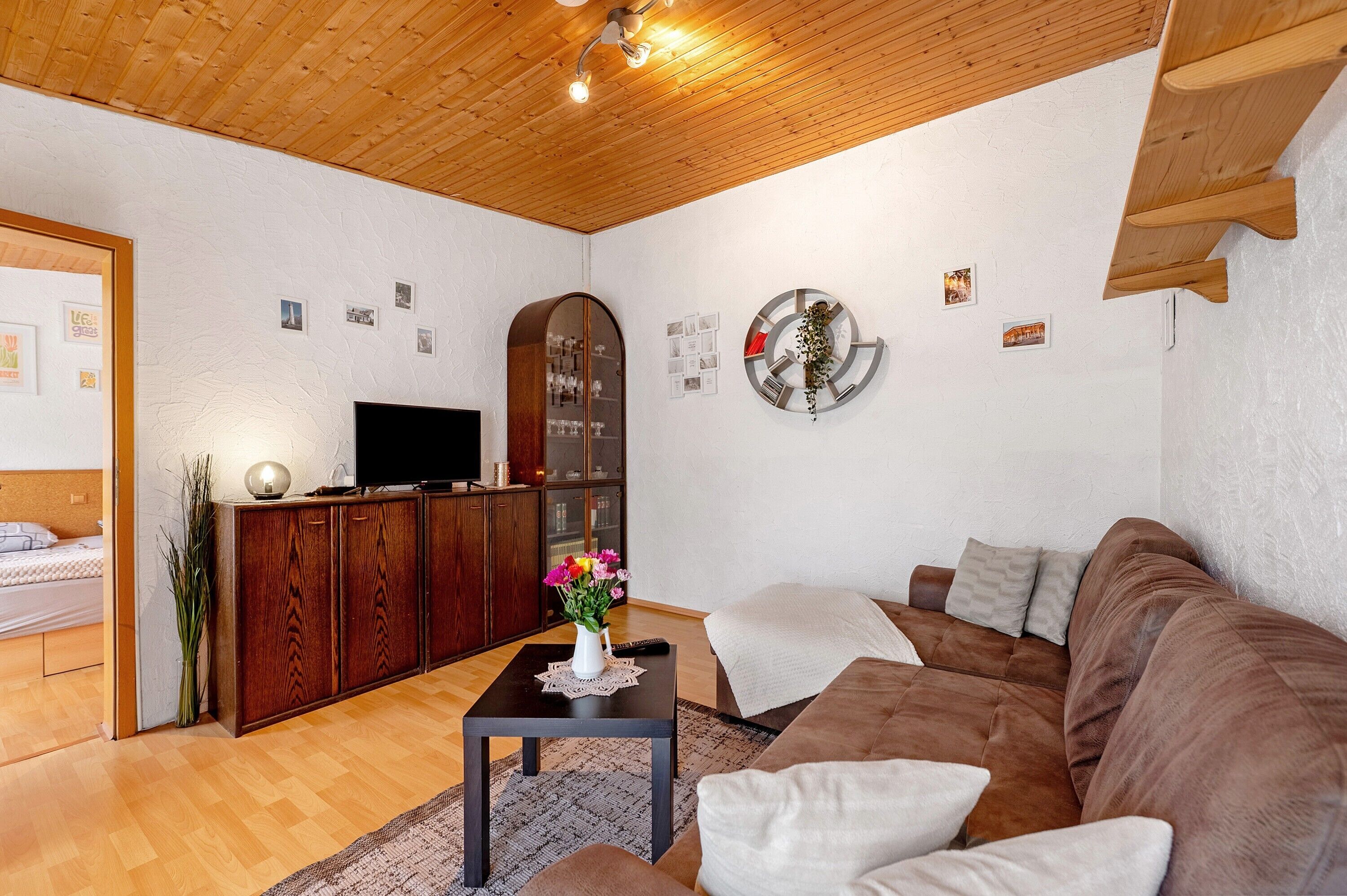 A living room with a wooden ceiling, a TV, a cabinet, a sofa, and a small table with a vase.