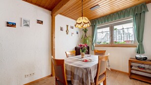 Ein Essbereich mit Holzboden, einem Tisch mit kariertem Tuch, Stühlen, einer Hängelampe und einem Fenster mit Vorhängen.