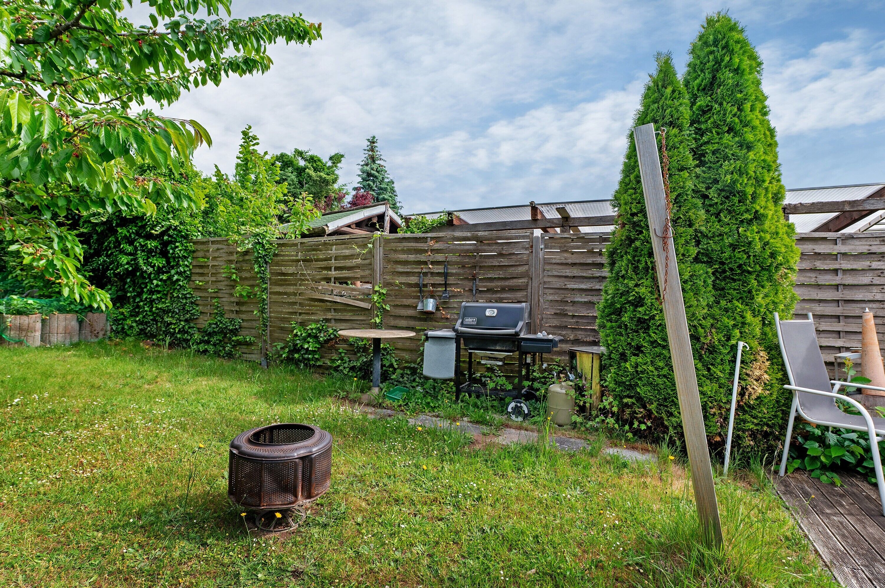Un patio trasero con valla de madera, una parrilla, una silla y un alto árbol de hoja perenne.