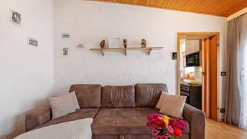 A living room with a brown sofa, a wooden ceiling, and a kitchen visible through an open door.