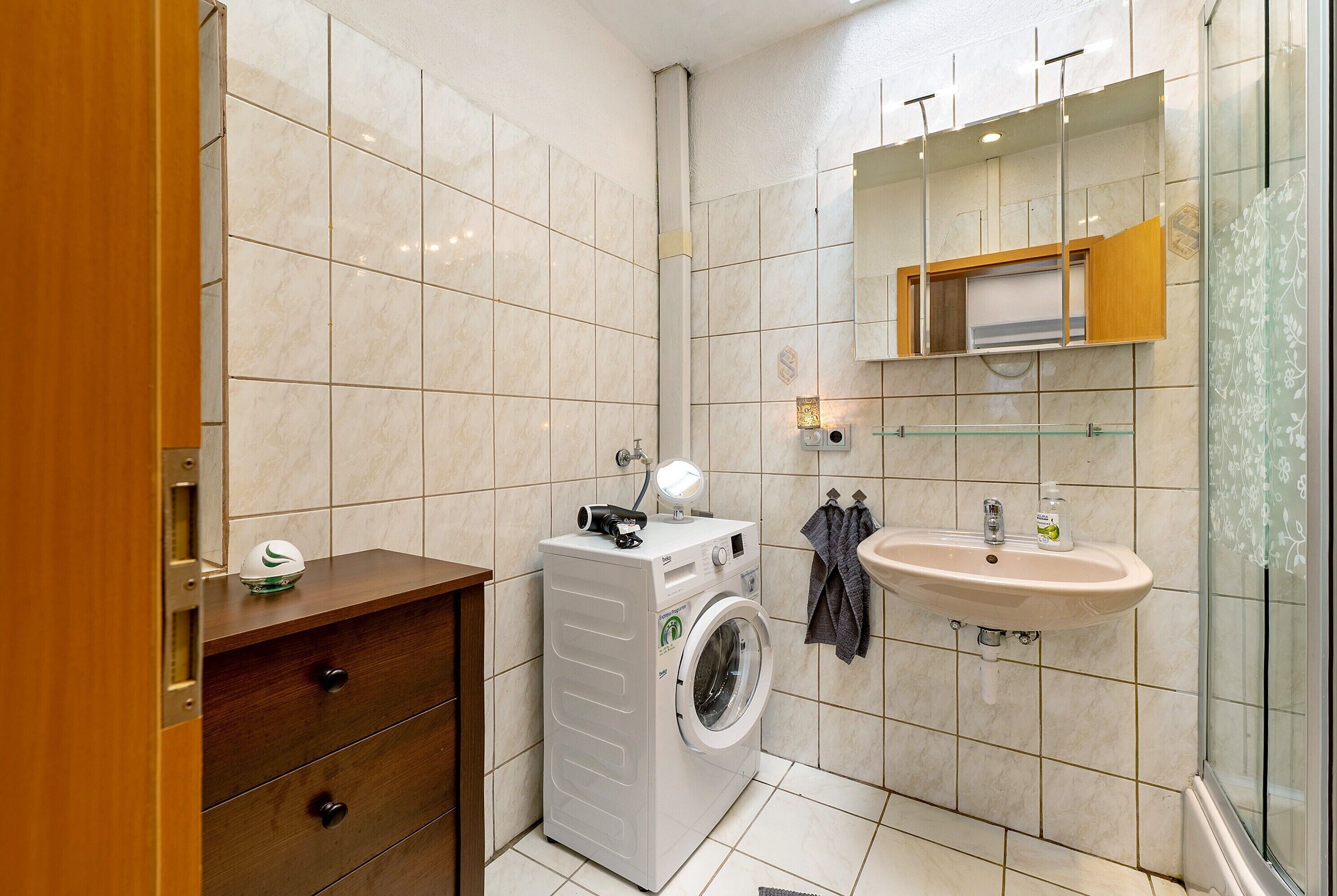 A compact bathroom with a washing machine, a sink, and a mirror.