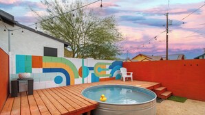 Ein Garten mit einer Holzterrasse, einem kleinen Pool mit einer Quietscheente, einem farbenfrohen Wandbild und einem weißen Stuhl.