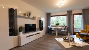 Ein modernes Wohnzimmer mit Flachbildfernseher, einem Essbereich mit Tisch und Stühlen sowie einem großen Fenster mit Vorhängen.