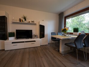 Ein modernes Wohnzimmer mit Flachbildfernseher, einem Esstisch mit Stühlen, einer Einbauküche und einem Fenster mit Blick auf Bäume.