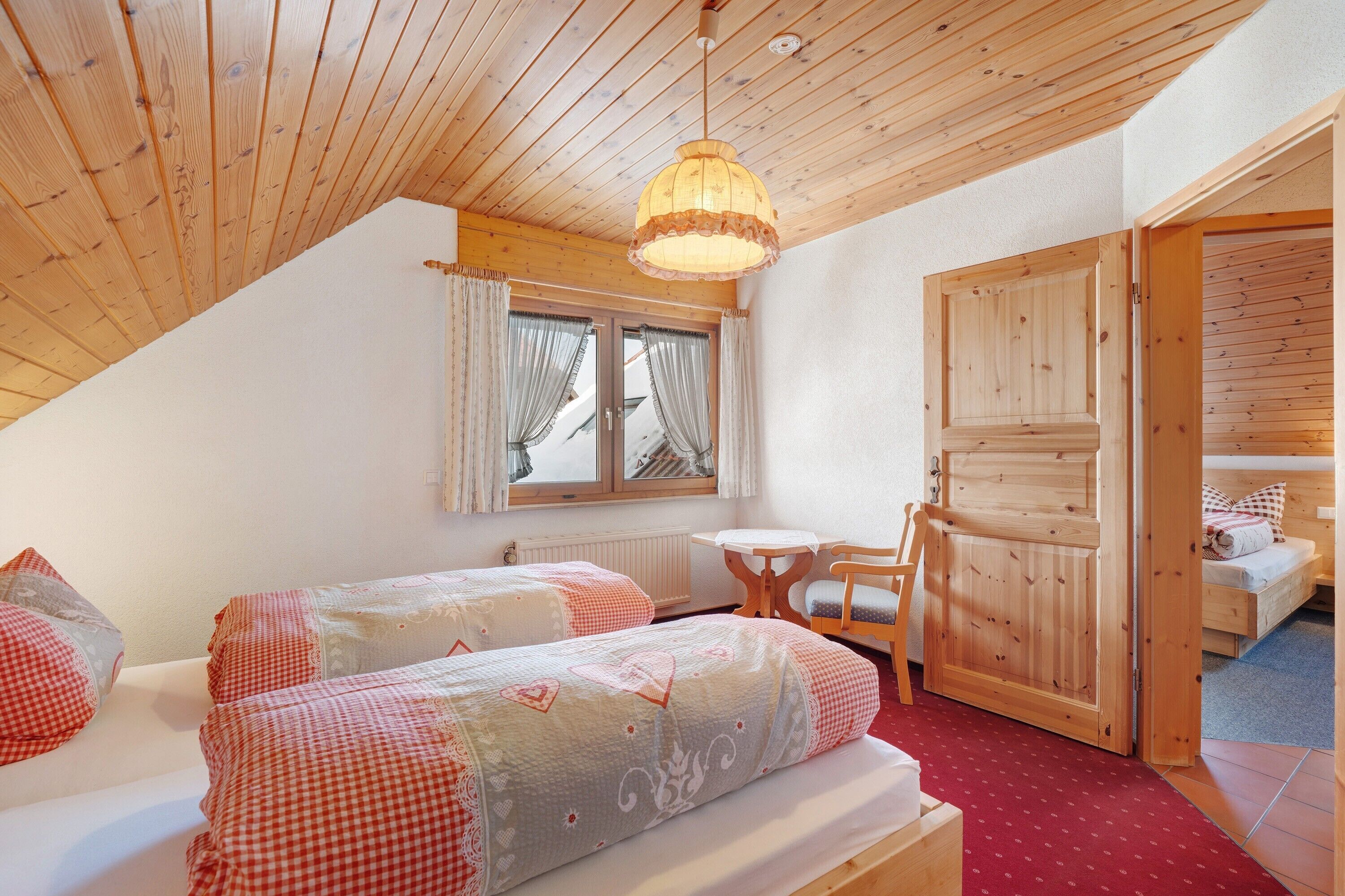 A cozy room with two beds, a wooden ceiling, a window with curtains, and a small table.