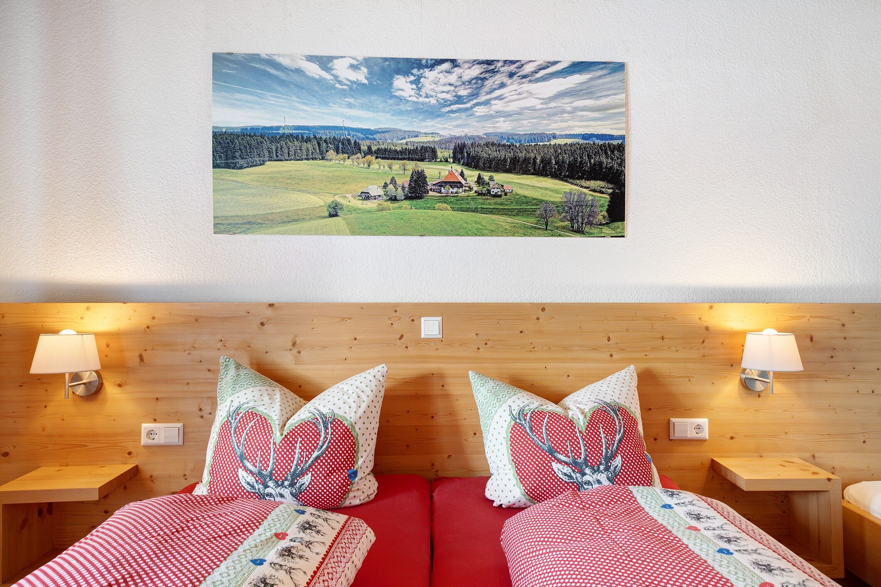 A room with a bed, two bedside tables, a wall-mounted lamp, and a framed landscape painting.