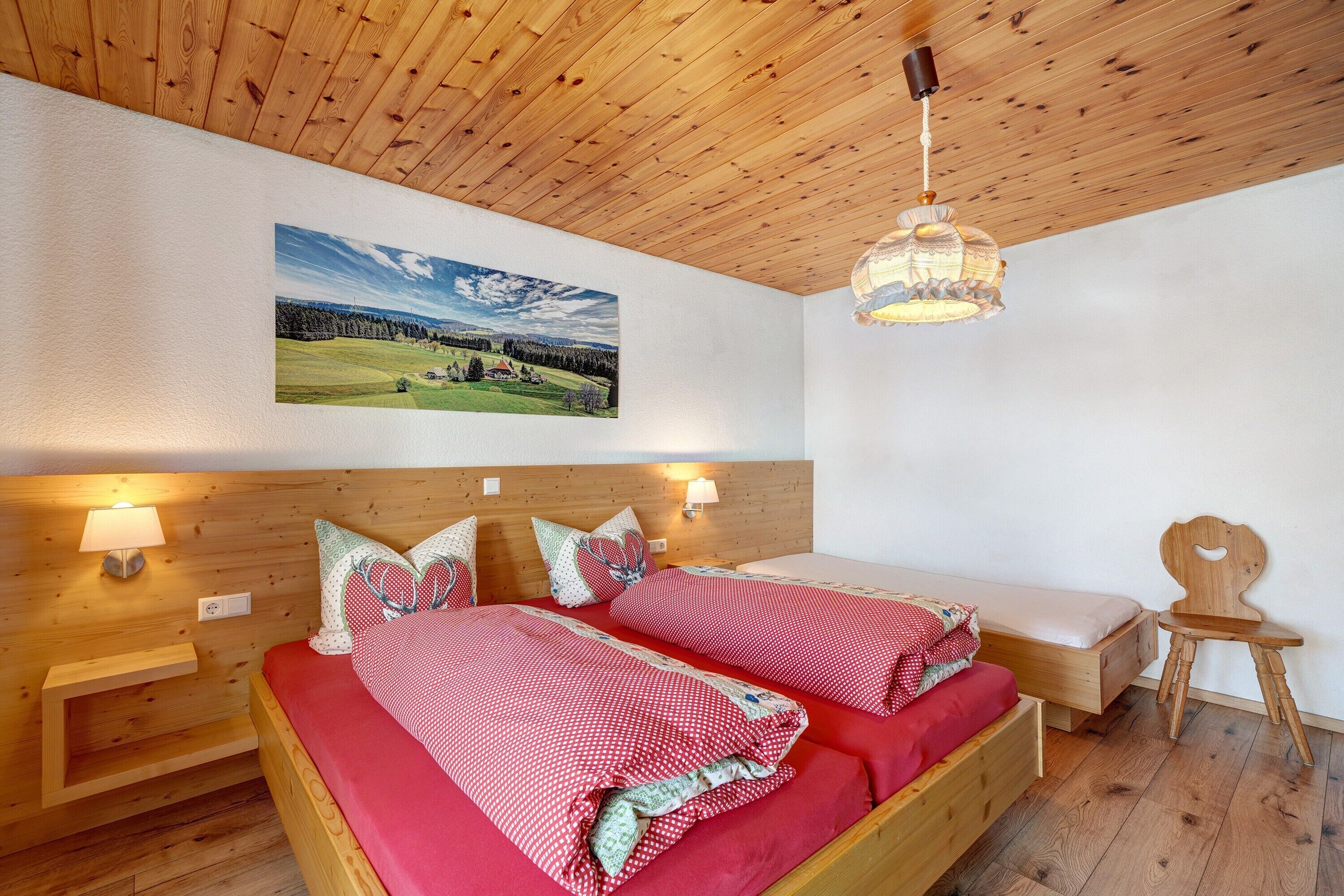A wooden ceiling, a bed with red bedding, a wooden nightstand, a wall-mounted lamp, and a landscape painting.
