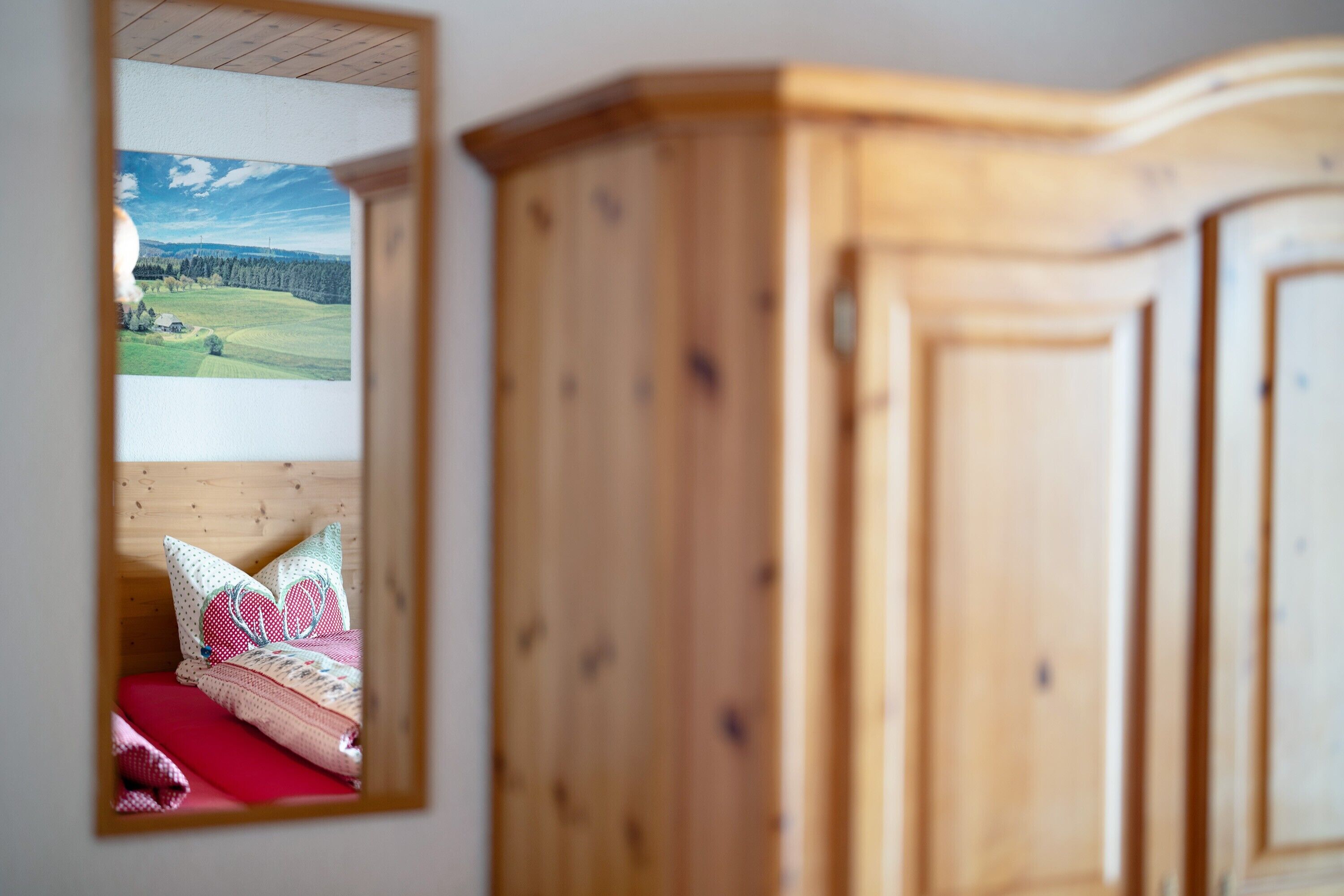A bedroom with a wooden wardrobe, a mirror reflecting a bed with a red bedspread and patterned pillows, and a framed landscape picture on the wall.