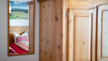 A bedroom with a wooden wardrobe, a mirror reflecting a bed with a red bedspread and patterned pillows, and a framed landscape picture on the wall.