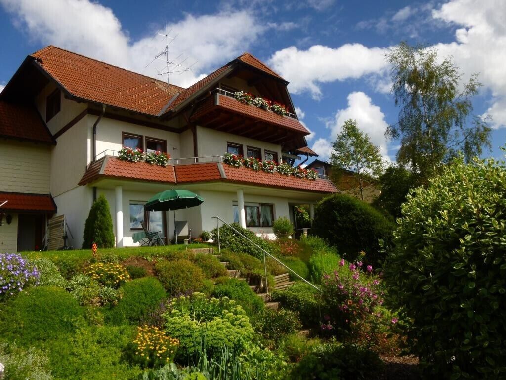 Une maison à deux étages, dotée d’un toit en pente, d’un balcon orné de fleurs et d’un jardin où poussent différentes plantes et fleurs.
