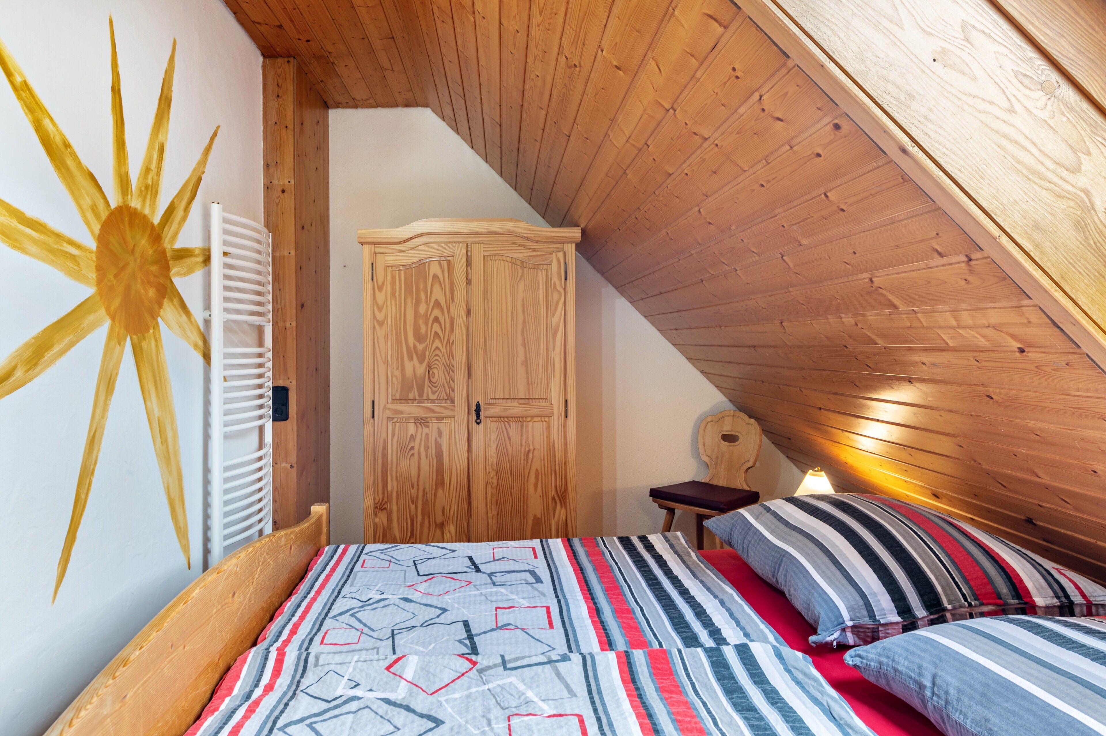 A bedroom with a sloped ceiling, a wooden wardrobe, a bed with striped bedding, and a wall with a sun design.