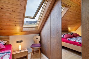 A bedroom with a sloped ceiling, a skylight, a bed with red bedding, a chair, and a bedside table with a lamp.