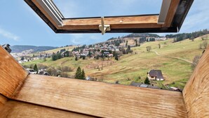 Une vue depuis une fenêtre à cadre en bois, donnant sur un paysage rural avec des maisons et des collines ondulantes.